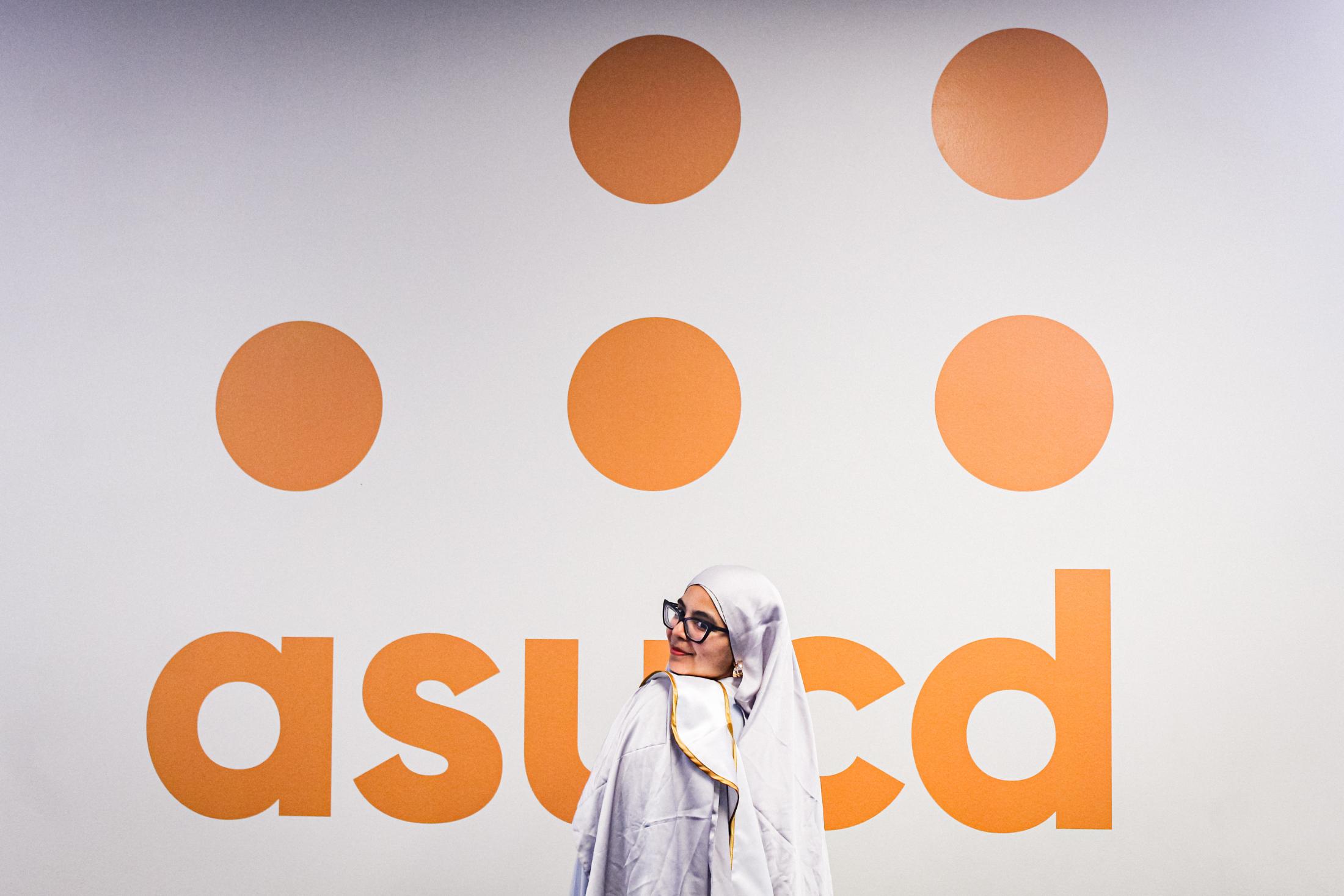 Aridin stands in a white hijab, outfit and graduation sash, in front of an orange Associated Students, University of California, Davis logo against a white background.