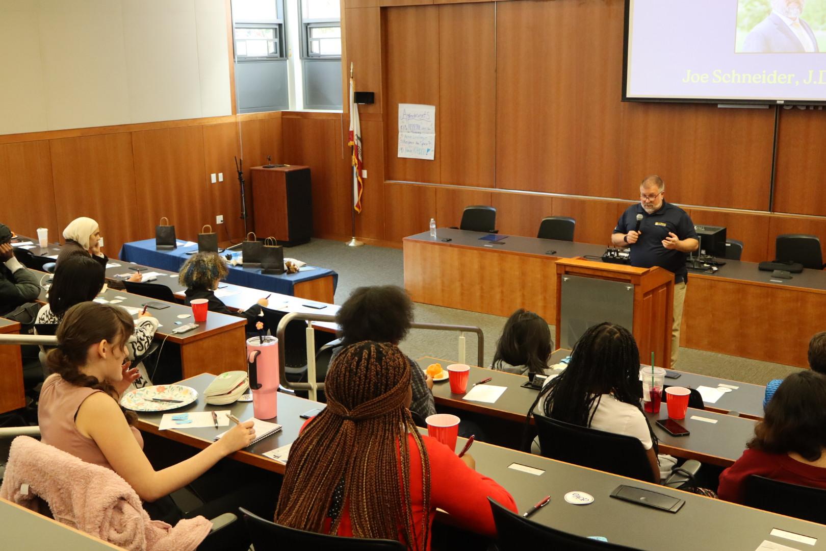 A UC Davis law school lecture hall filled with high school students attentively listening to a speaker at a podium. The speaker, Joe Schneider, stands at the front with a microphone, giving a presentation about legal education during Discover Law Day.