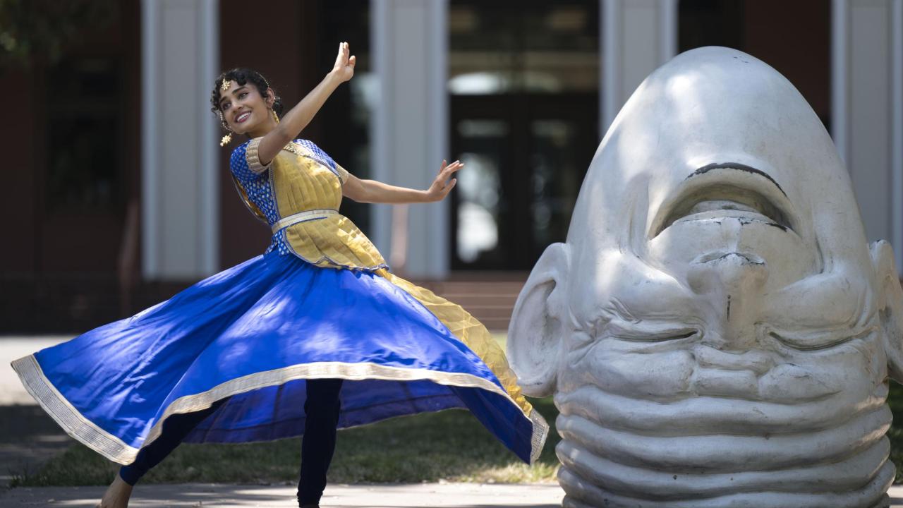 Vyoma, in a blue and gold traditional Kathak dance dress, spins next to the egghead sculpture, Eye on Mrak (Fatal Laff).