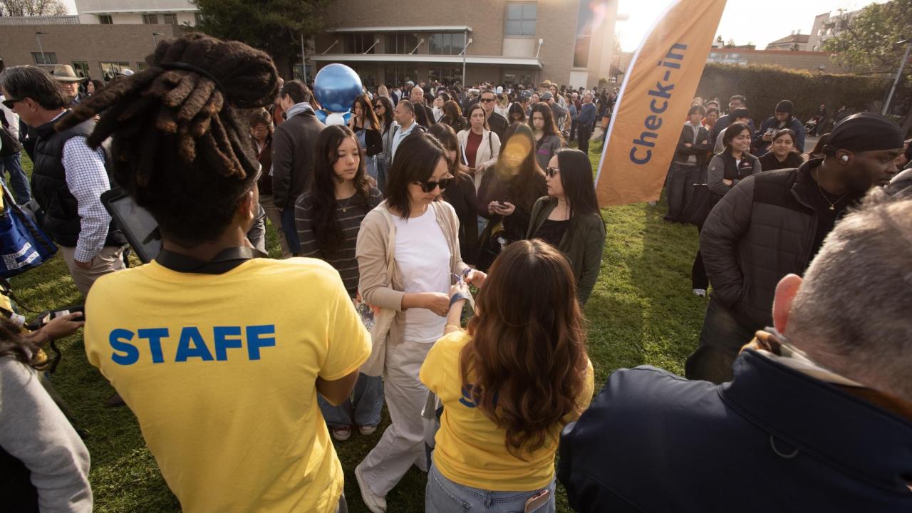 Admitted students and their families line up in small crowds at the Aggie Day check-in area, where staff in yellow t-shirts with blue writing that says staff help guide them into campus. 