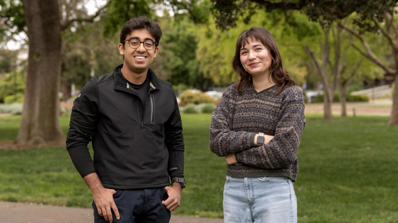 Two UC Davis students, both Goldwater Scholars, stand side by side outdoors on campus. The student on the left wears glasses, a black quarter-zip jacket and navy pants. The student on the right wears a multicolored sweater and light blue jeans. Both smile at the camera with trees and green grass in the background.