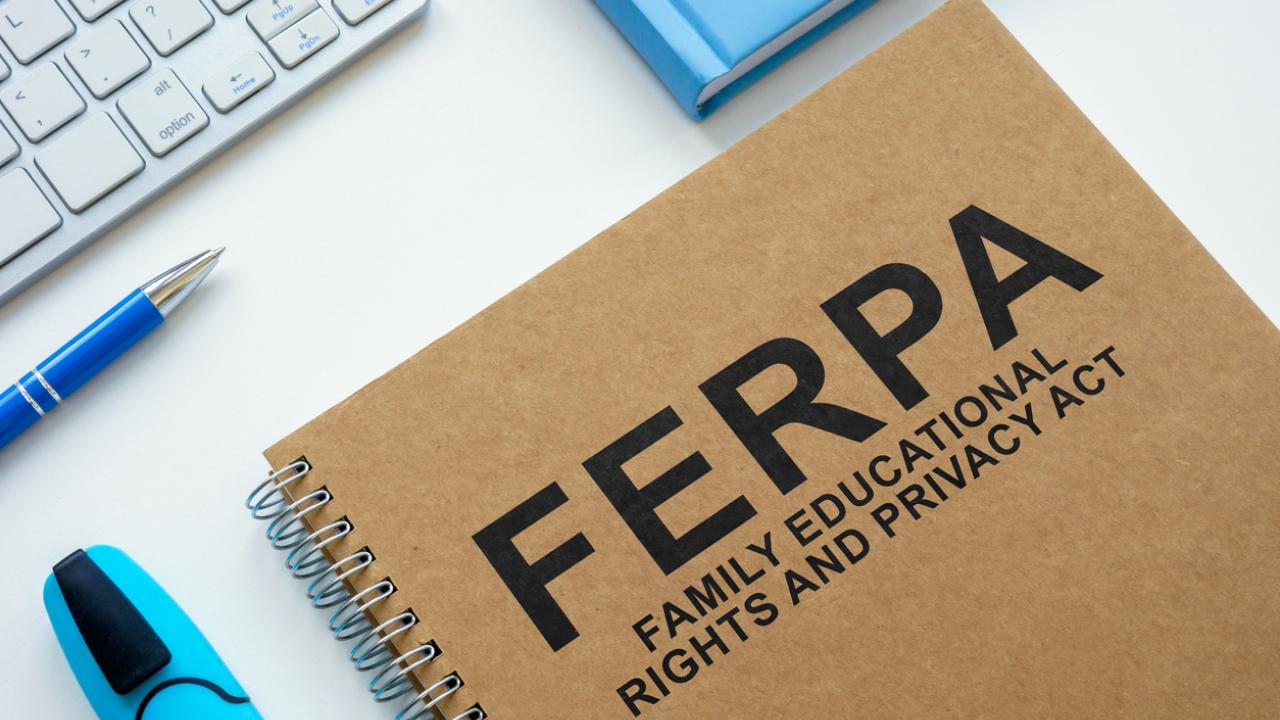 A notebook with FERPA Family Educational Rights and Privacy Act imprinted on its cover rests on top of the keyboard of an open laptop computer against an aqua blue background.
