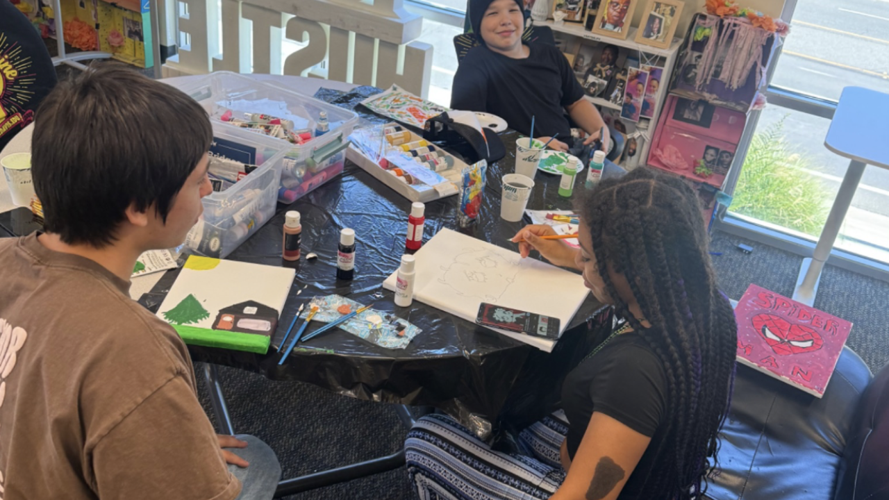 Three students paint canvases together at a table covered with art supplies.