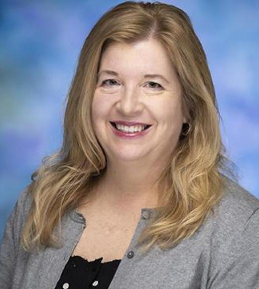 Studio headshot photograph of smiling blonde woman in gray cardigan against blurred blue background