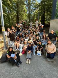 A large group of students and staff smile and pose together on outdoor steps surrounded by trees.