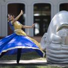 Vyoma, in a blue and gold traditional Kathak dance dress, spins next to the egghead sculpture, Eye on Mrak (Fatal Laff).