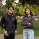 Two UC Davis students, both Goldwater Scholars, stand side by side outdoors on campus. The student on the left wears glasses, a black quarter-zip jacket and navy pants. The student on the right wears a multicolored sweater and light blue jeans. Both smile at the camera with trees and green grass in the background.