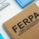 A notebook with FERPA Family Educational Rights and Privacy Act imprinted on its cover rests on top of the keyboard of an open laptop computer against an aqua blue background.