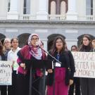 Aridin speaks at a podium in a hot pink outfit in front of a government building, surrounded by colleagues with signs that read “Education shouldn’t be a privilege” and “Budget better have my money.”
