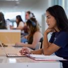 A student sits at a desk and works on a laptop with an open binder in front of them in a study space with other students in the background.