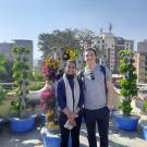 Two people stand on a rooftop garden surrounded by vertical hydroponic towers filled with leafy greens and flowers, with city buildings in the background on a sunny day.